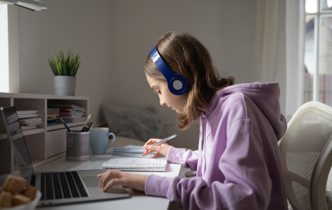 A person wearing blue headphones and a purple hoodie sits at a desk, writing in a notebook with a laptop, plant, and coffee mug nearby.