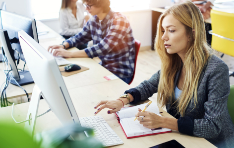 A woman works at a computer while taking notes in a notebook at a shared office desk with other people working in the background.