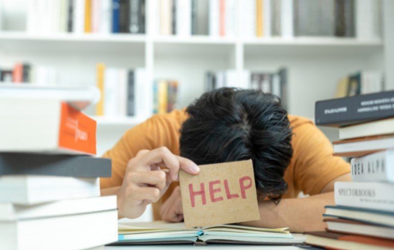 A person sits at a desk with their head down, surrounded by stacks of books, holding a cardboard sign that reads "HELP.