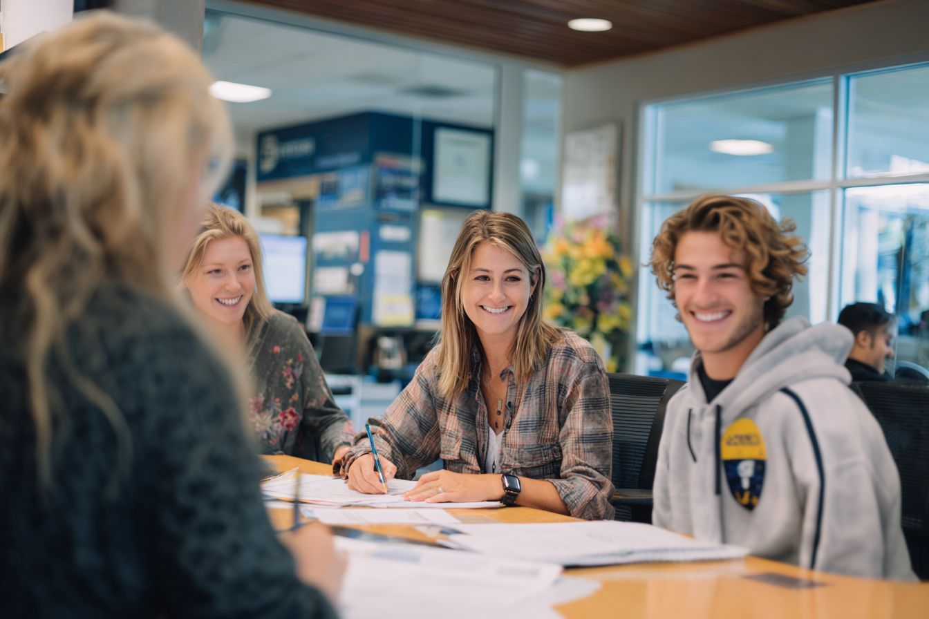 Three young adults sit at a table with papers, smiling and talking, in a brightly lit office or school setting.