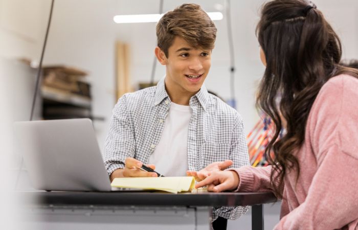 Two teenagers sit at a table with a laptop and notebook, having a conversation. One is writing while the other listens.