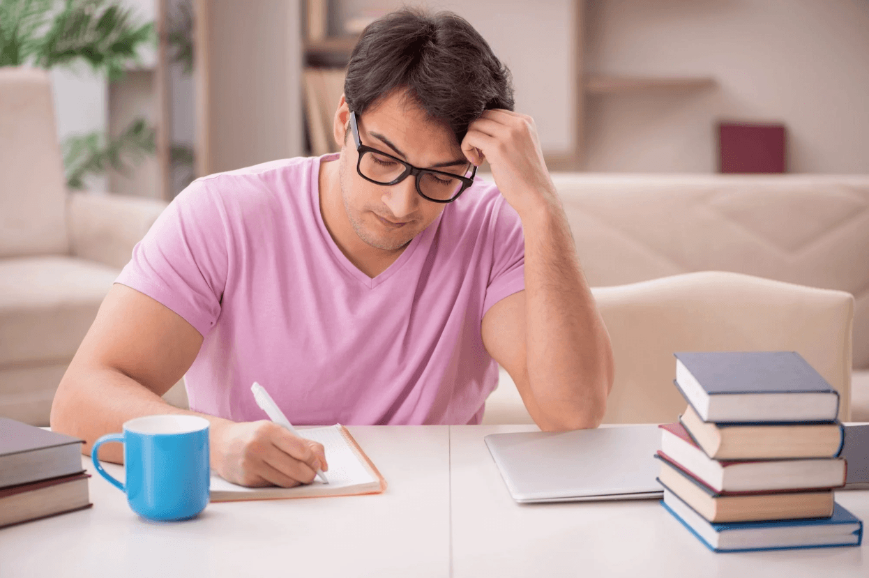 A young man in a pink shirt writing in a notebook with a thoughtful expression.