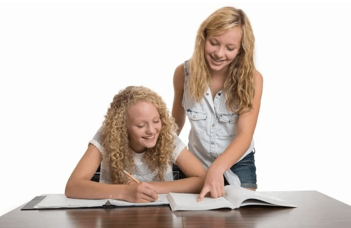 Two girls studying together at a table, one writing and the other pointing at a book.